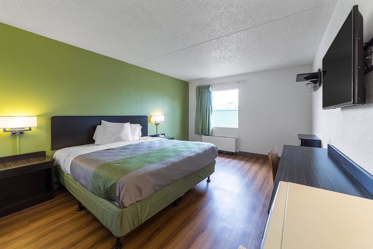 A hotel room with a bed, bedside tables, a desk, a chair, a window with curtains, a flat-screen TV mounted on the wall, and a radiator under the window.