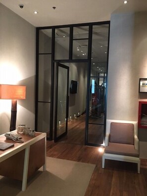 Quarto de hotel moderno com piso de madeira, uma escrivaninha com abajur, uma cadeira e um espelho grande.