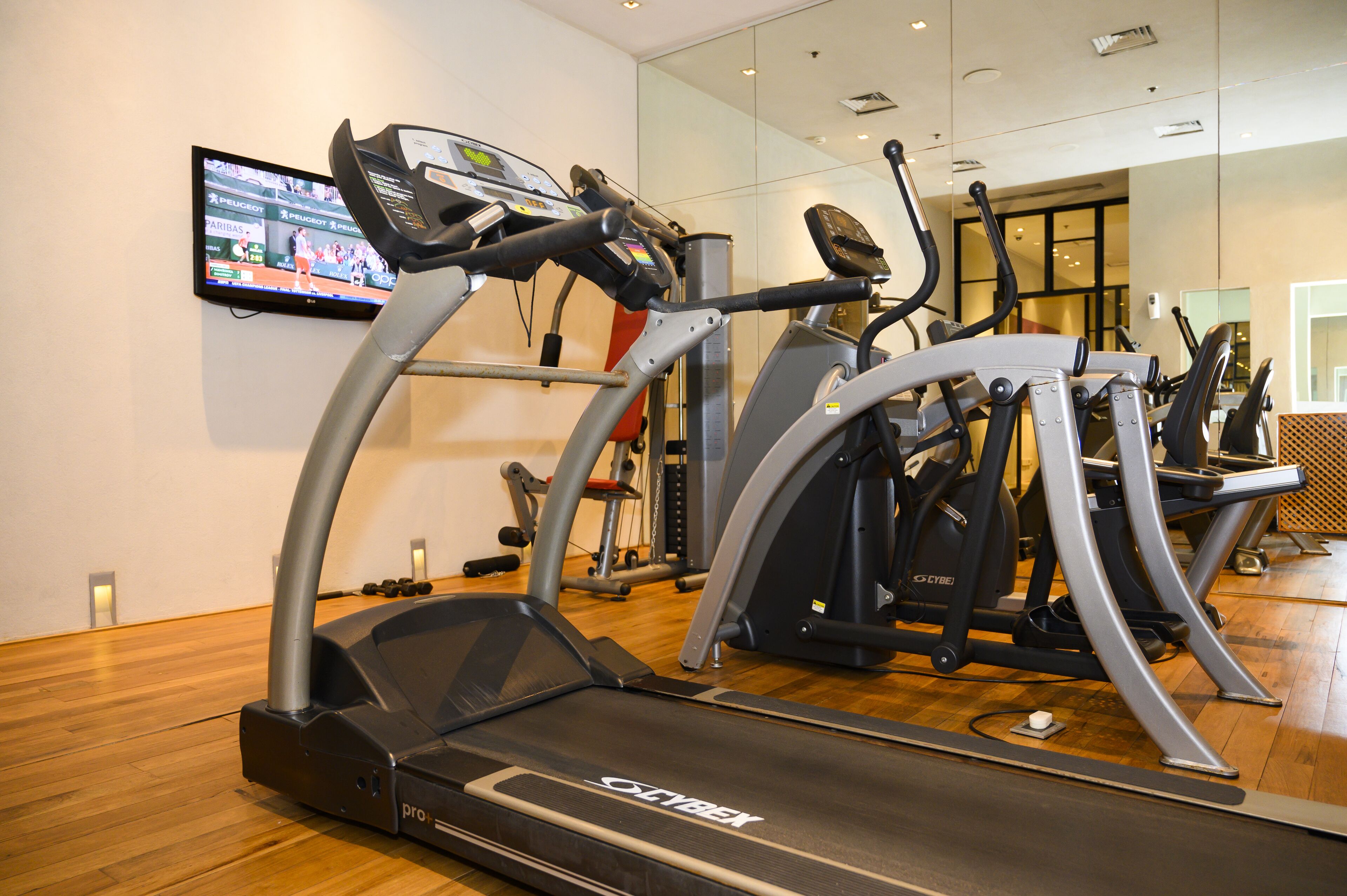 Un gimnasio con cintas de correr, elípticas y un televisor mostrando deportes.