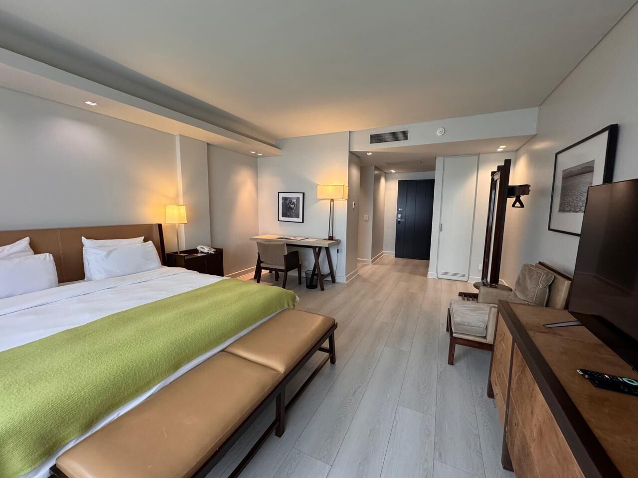 Ein Hotelzimmer mit Bett, Schreibtisch, Stuhl, Bank, Fernseher und einer Tür zu einem weiteren Raum.