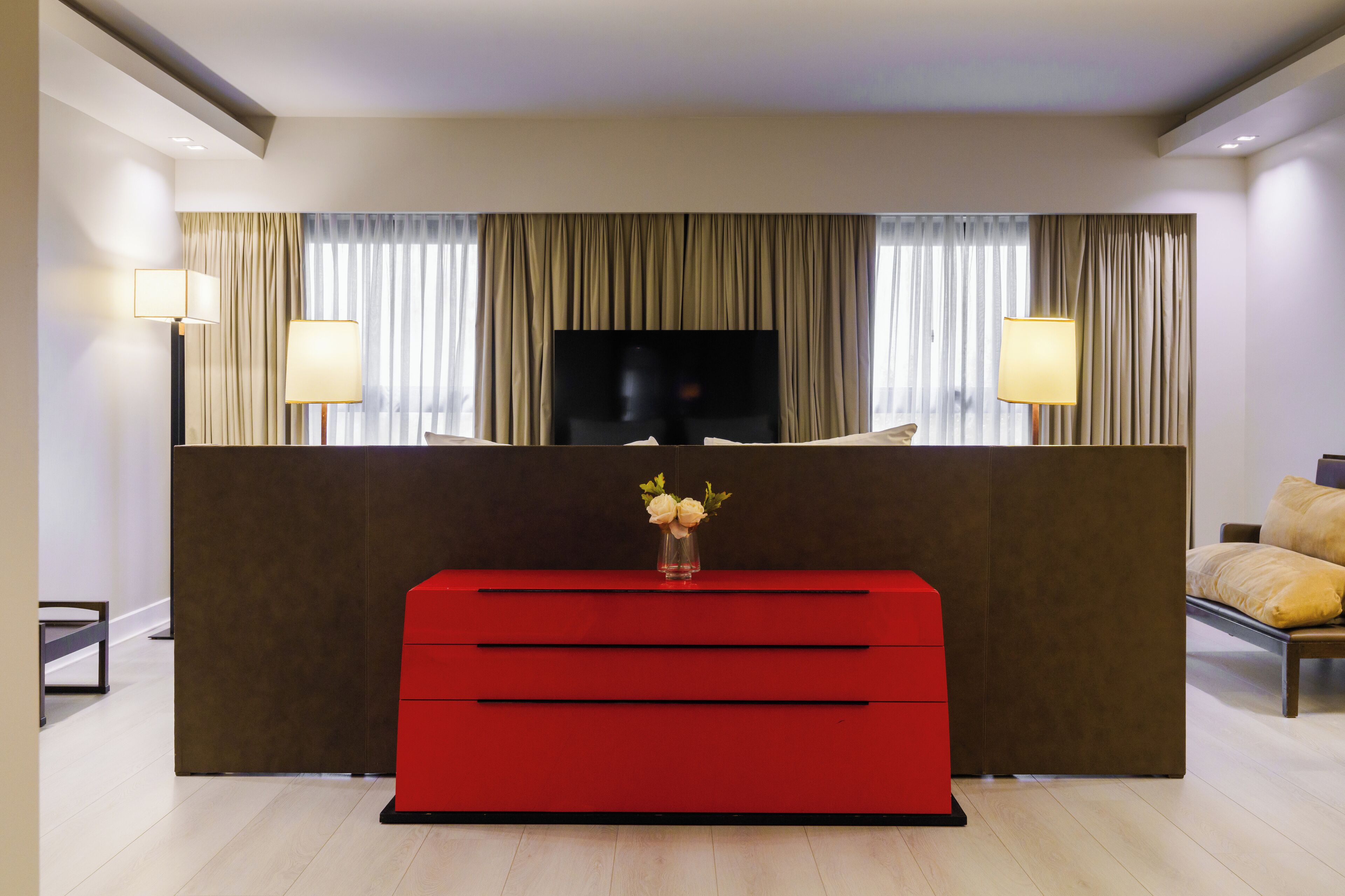 Una habitación de hotel moderna con un televisor de pantalla plana grande, una mesa consola roja con un florero y una cama con almohadas color beige.