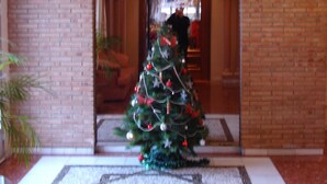 Un árbol de Navidad adornado con adornos y luces en un área de recepción con suelo de baldosas y paredes de ladrillo.