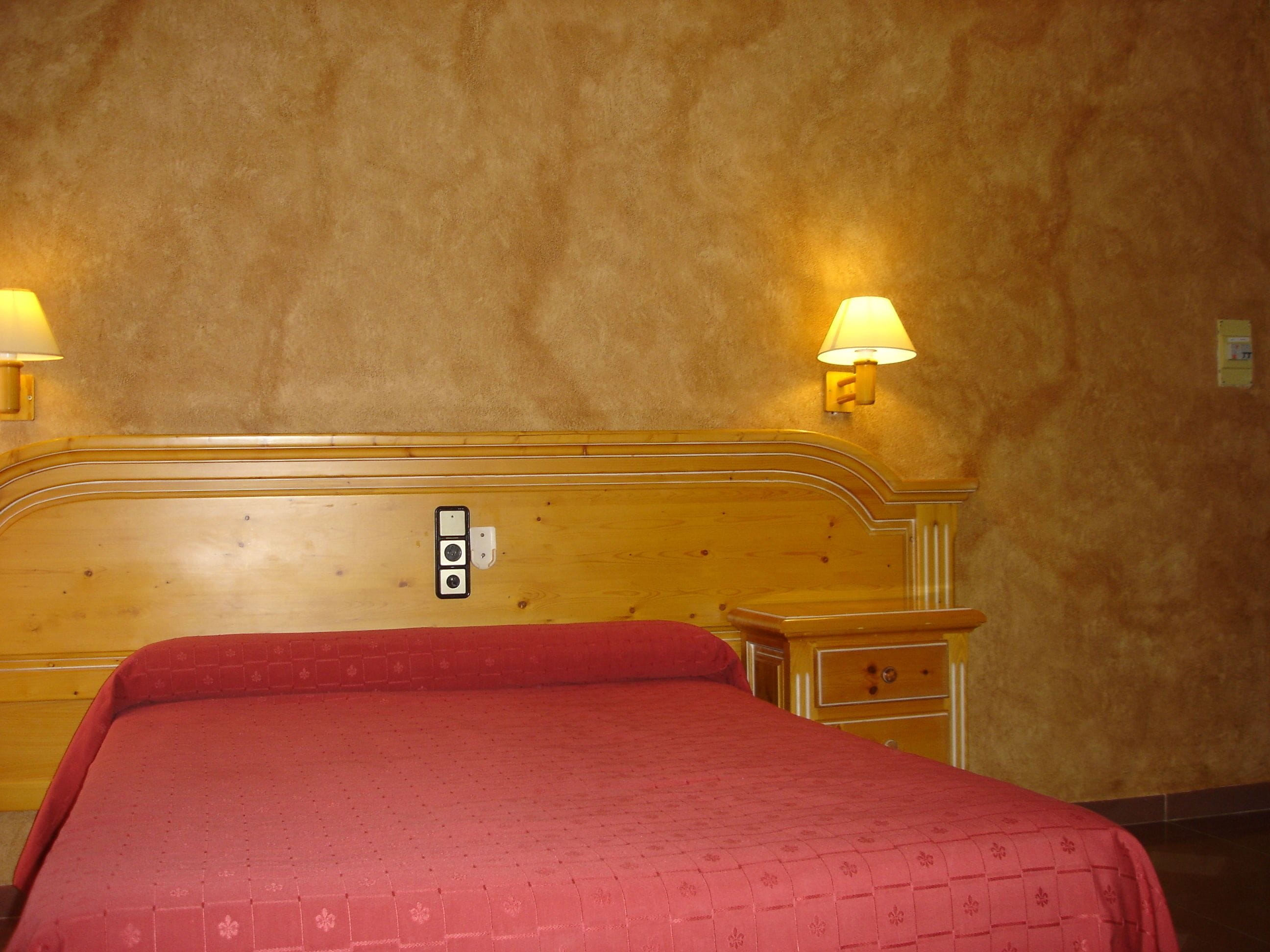 Habitació d'hotel amb un panell de capçal de fusta, una colcha vermella, dues làmpades de nit i una tauleta de nit amb calaixos.