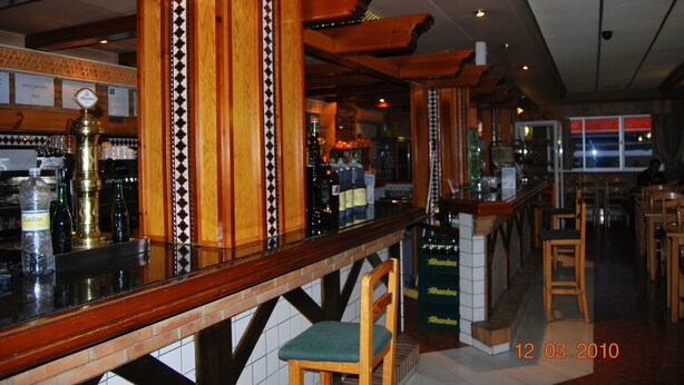 Una zona de bar con vigas de madera, barra y taburetes. Hay botellas y vasos sobre la barra.