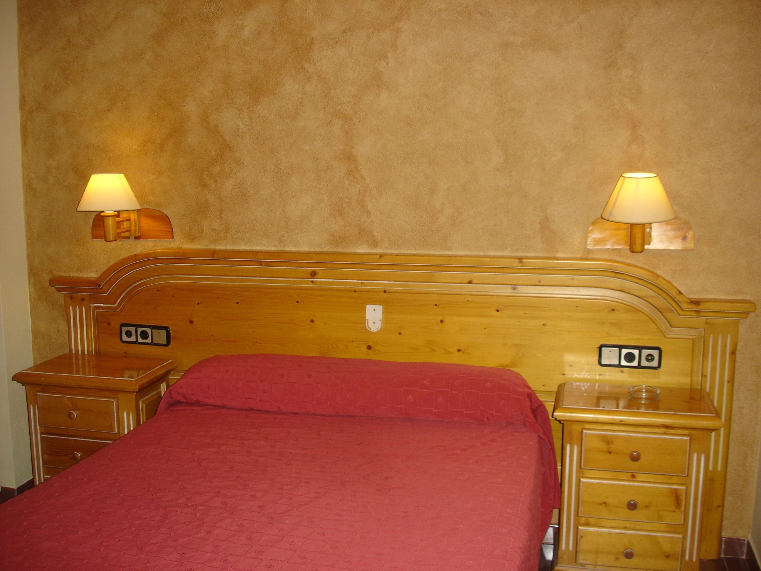 A hotel room with a wooden headboard, a red bedspread, two bedside tables with lamps, and a small wooden nightstand with drawers.