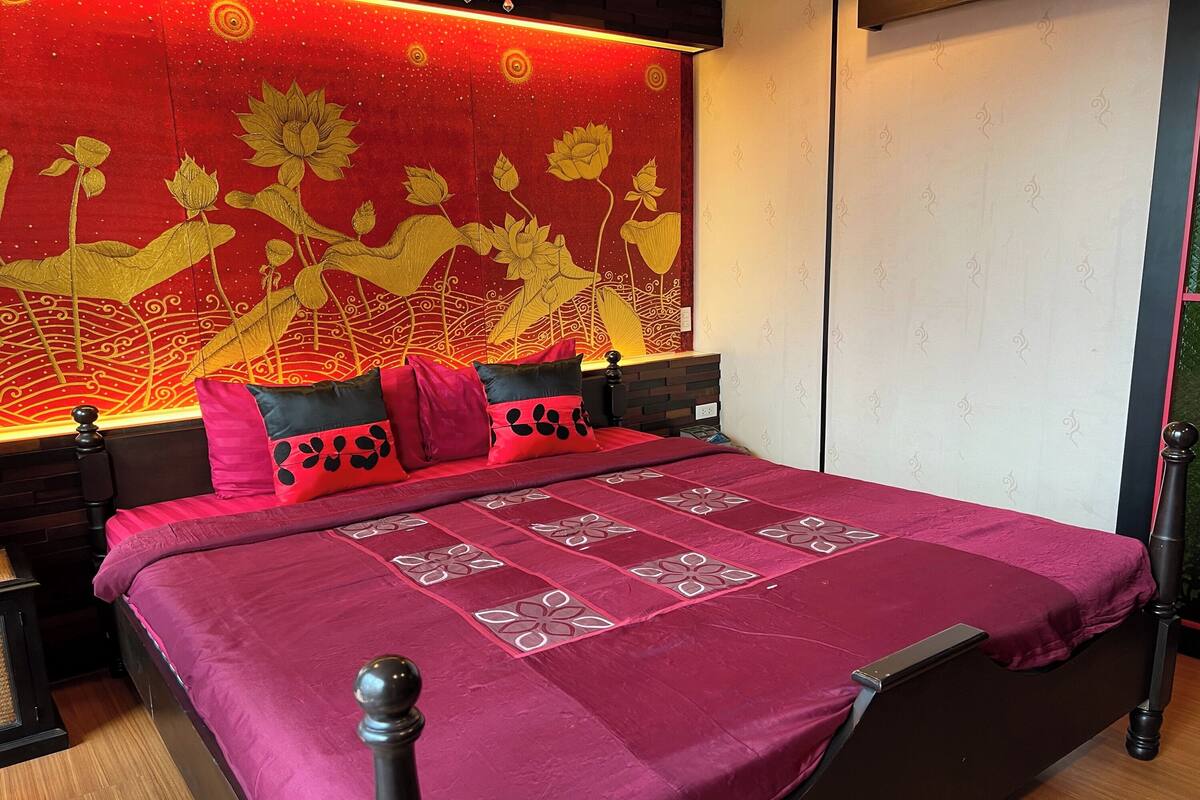 Un lit recouvert d’une couvre-lit aux motifs rouges et noirs, une grande tapisserie murale représentant des fleurs de lotus et une table de chevet avec une lampe.