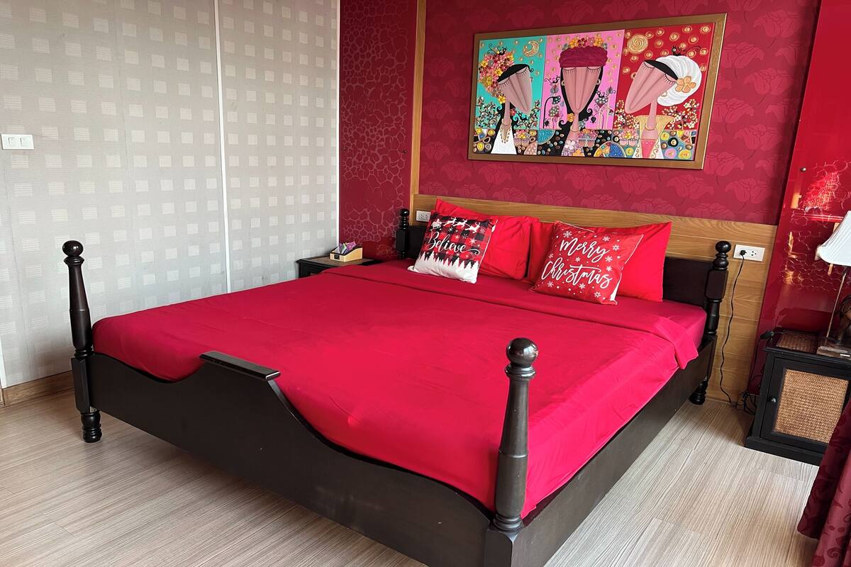 Une chambre à coucher avec une couvre-lit rouge, une tête de lit en bois et un tableau coloré accroché au mur.