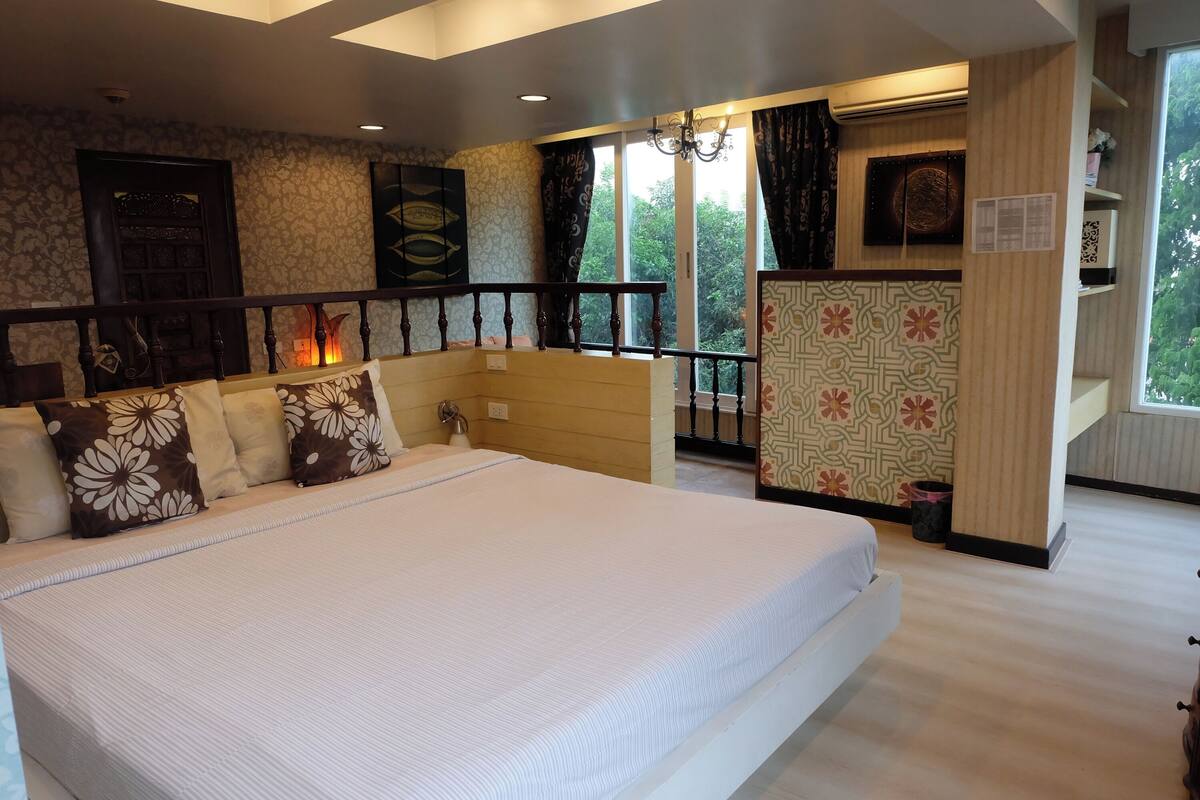 Une chambre à coucher avec un grand lit, une tête de lit en bois, une petite table de chevet, une commode, une fenêtre avec des rideaux et une vue sur les arbres à l’extérieur.