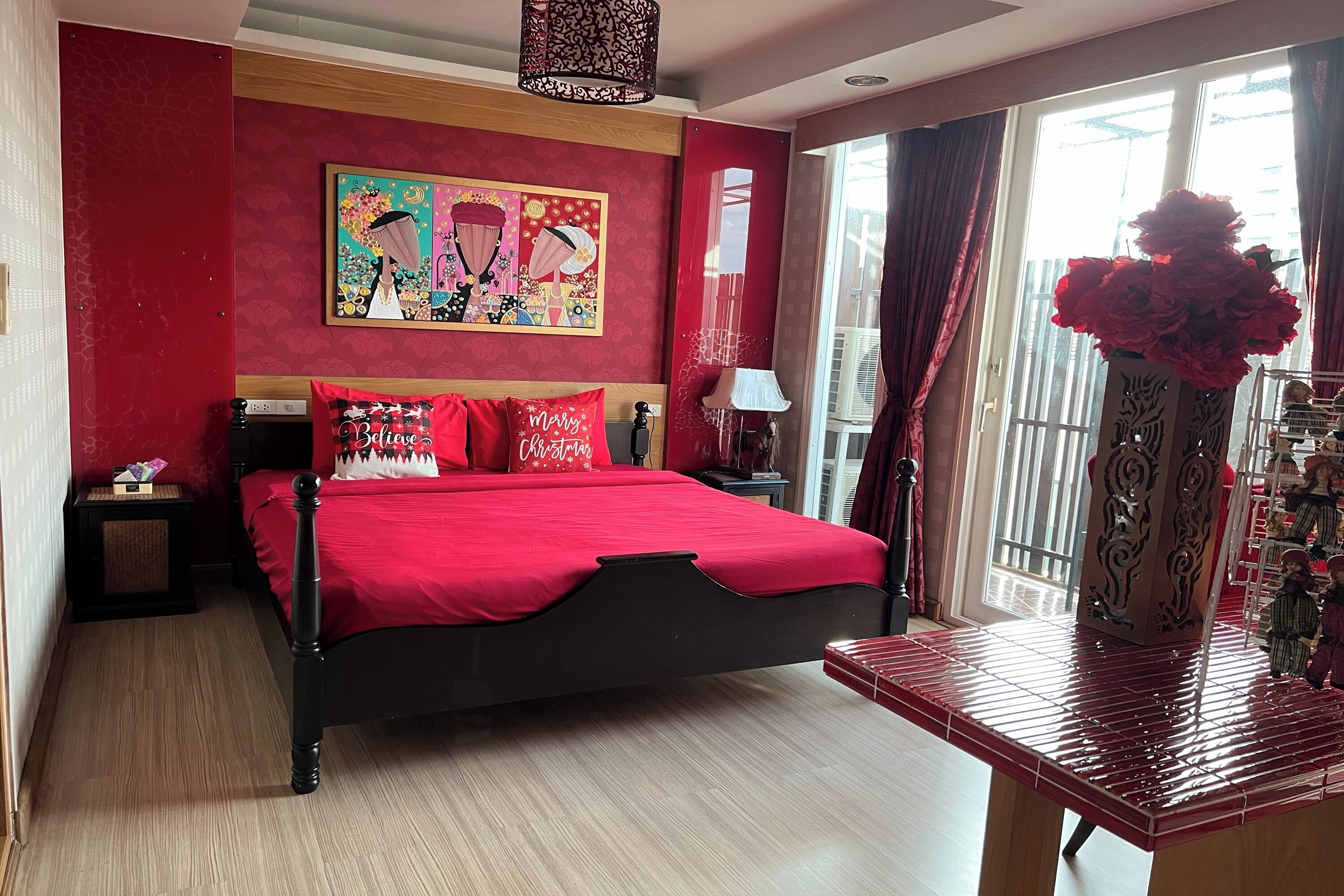 Un dormitorio con una colcha roja, una mesita de noche, una lámpara, un florero decorativo y un cuadro en la pared.