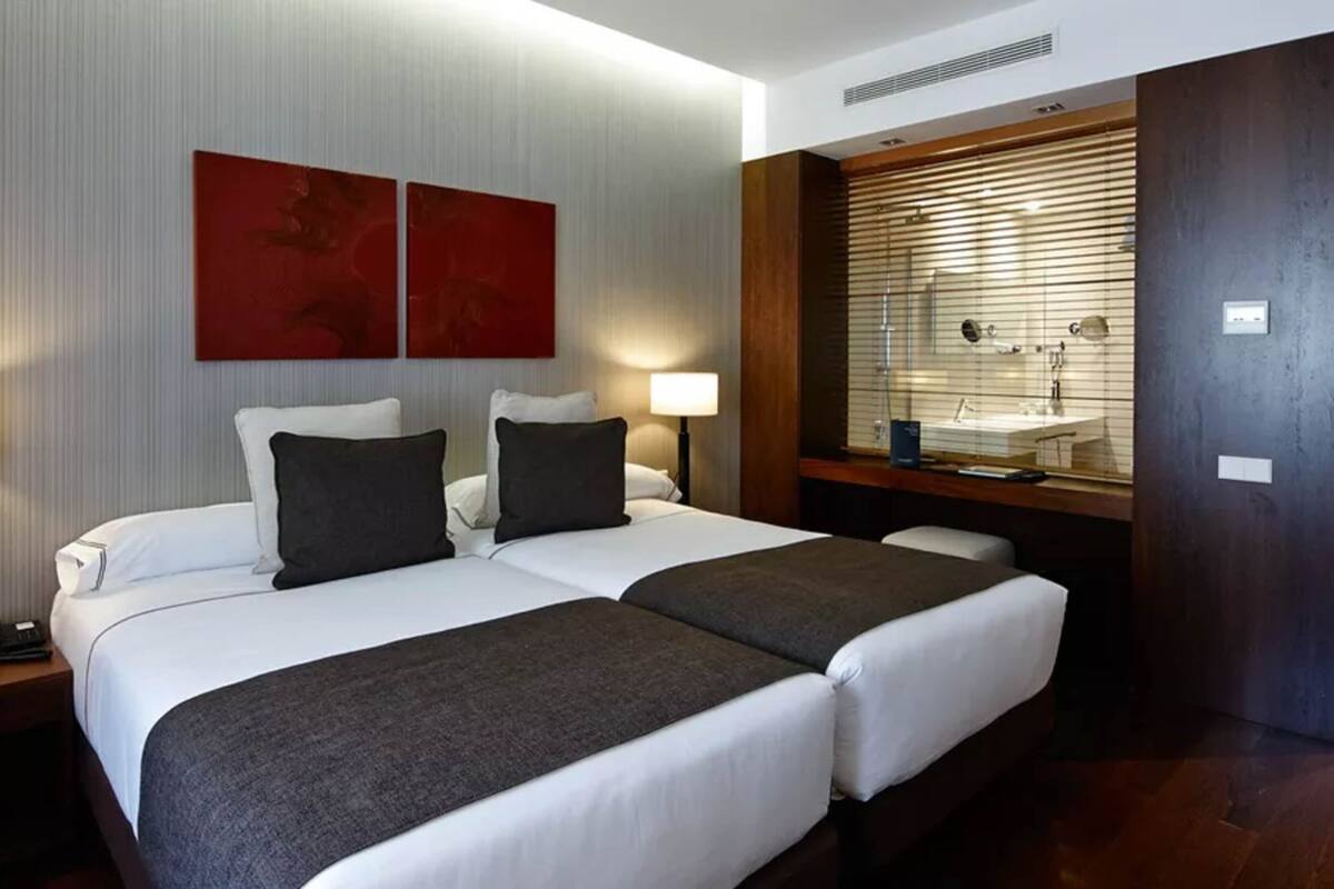 A hotel room with two beds, a wooden bench, a vanity with a mirror, and a red abstract painting on the wall.