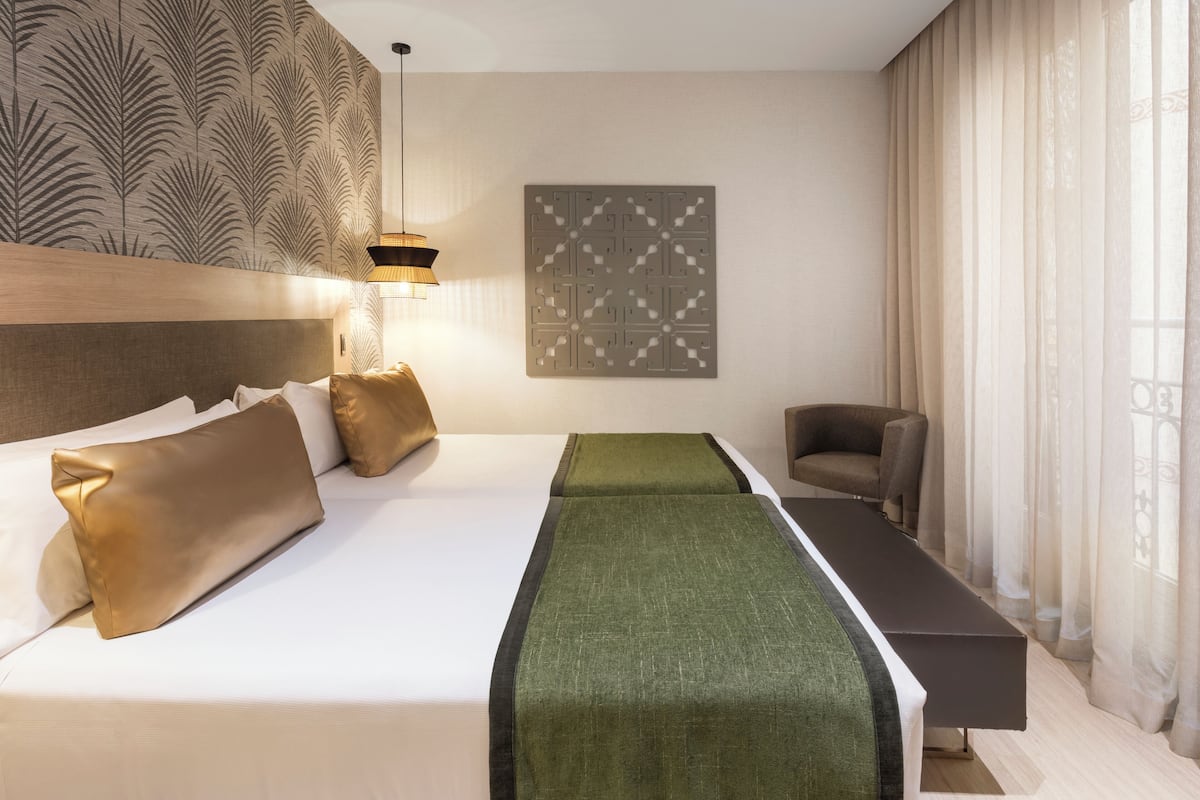 A hotel room with a large bed, a bench, a chair, a wall-mounted artwork, and a patterned wallpaper.