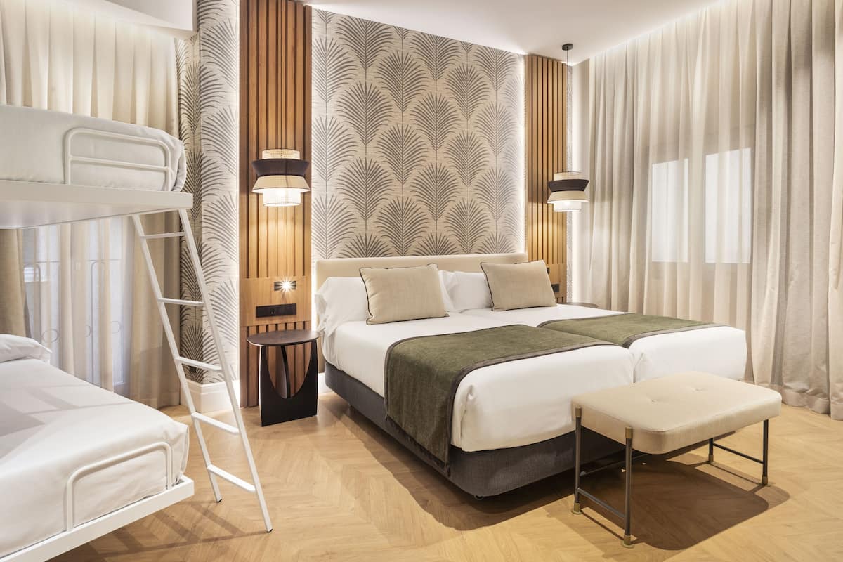 A modern hotel room with a bunk bed, a large bed with a green throw, wooden paneling on the wall, and two bedside lamps.