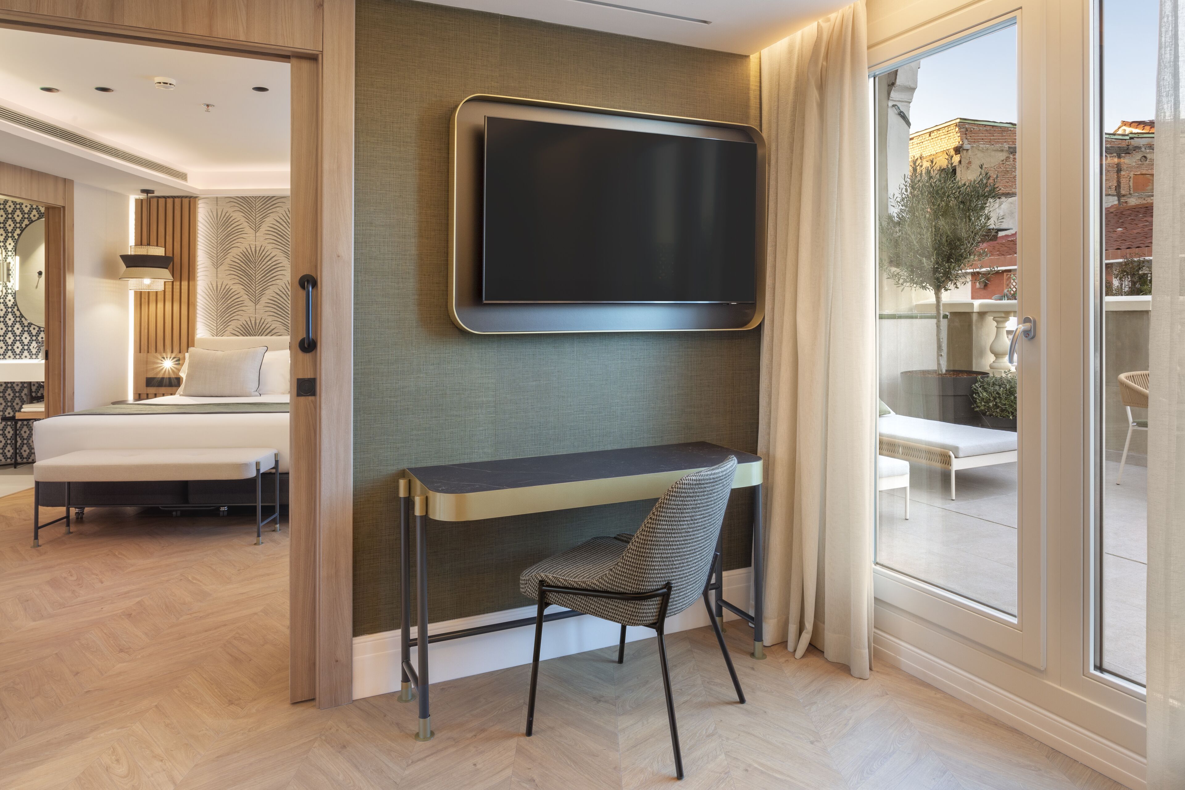 A modern hotel room with a large TV mounted on the wall, a desk with a chair, a bed, and a balcony with a view of the city.