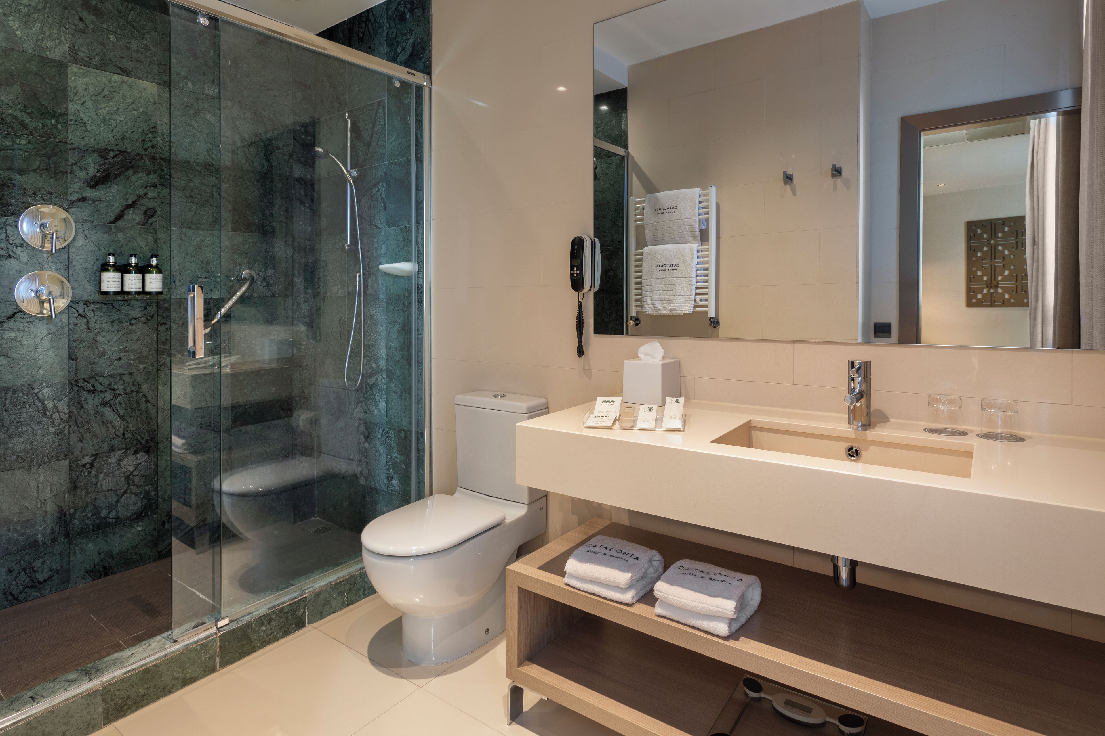 Un baño moderno con ducha envuelta en vidrio, inodoro, lavabo con espejo y repisa con toallas.