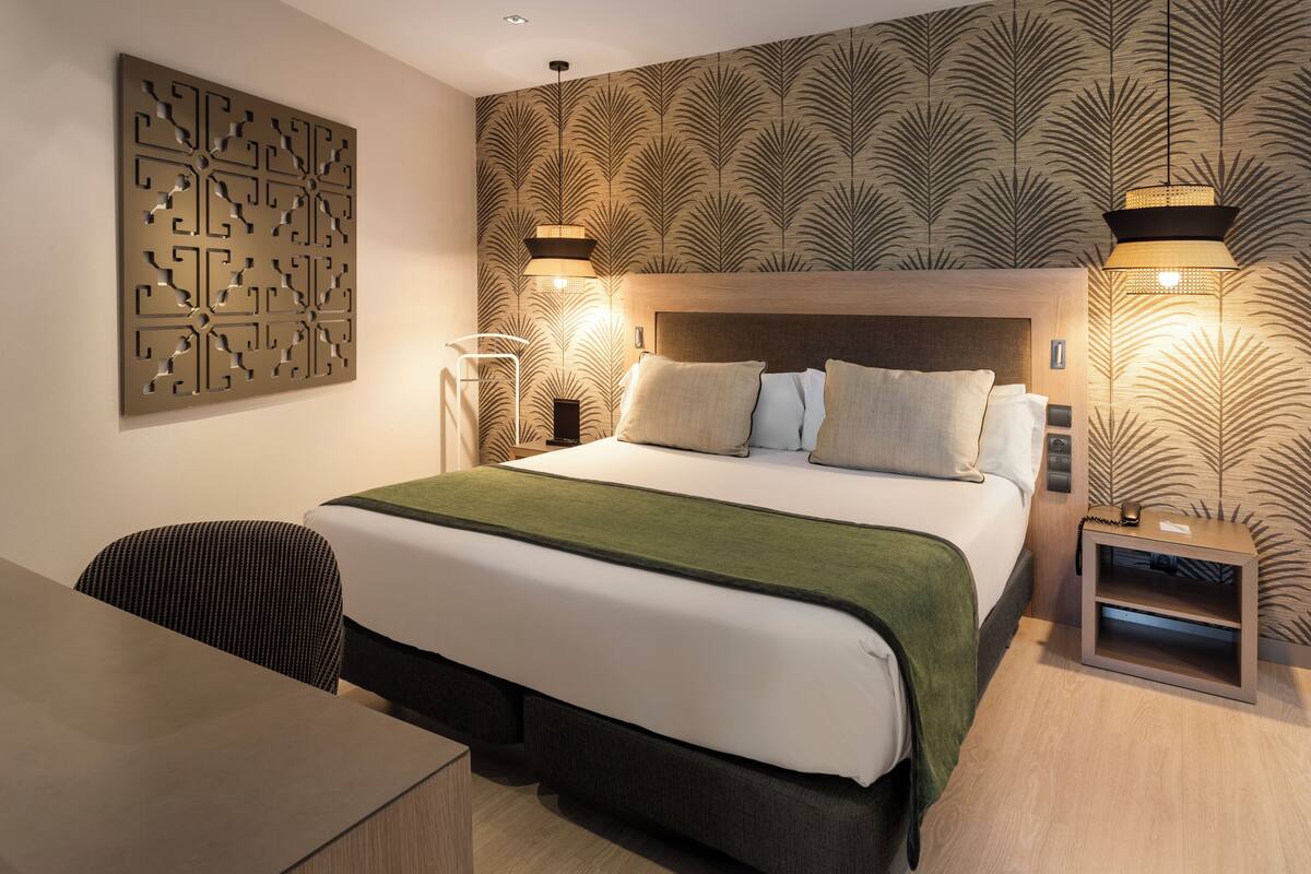 A hotel room with a large bed, a desk, and a chair. There are two wall-mounted lamps and a decorative wall panel.