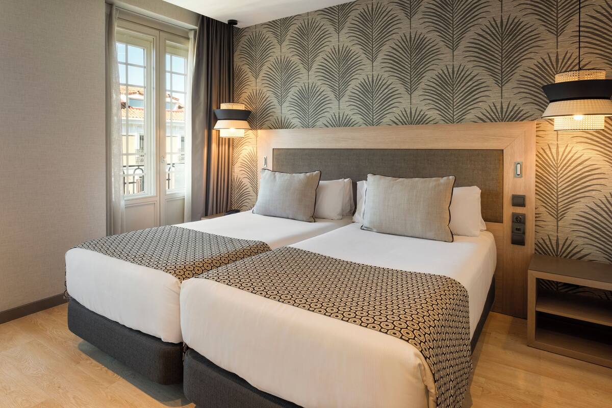 A hotel room with two beds, a large window, and a patterned wallpaper.