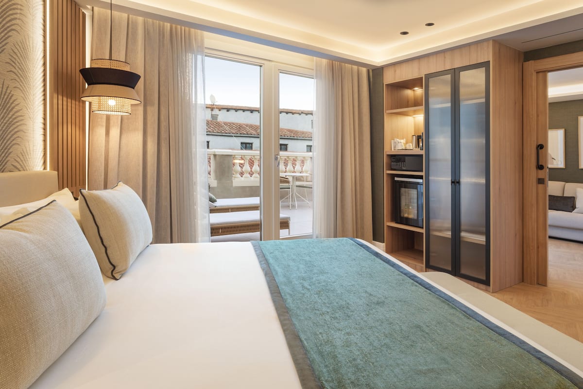 A hotel room with a bed, a desk, a wardrobe, and a balcony with a view of buildings.