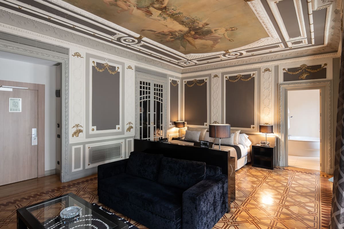 A luxurious hotel room with a classic design, featuring a large bed, a dark blue sofa, and a decorative ceiling.