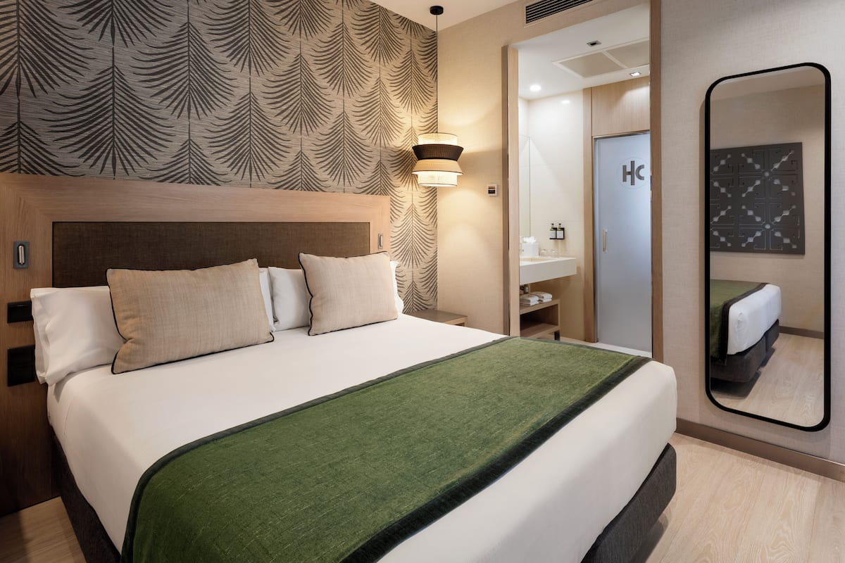 A hotel room with a large bed, a headboard, a green throw blanket, a wall-mounted mirror, and a patterned wallpaper.
