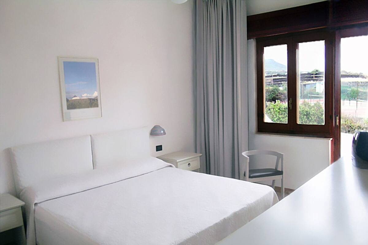 A hotel room with a bed, a nightstand, a chair, a window with a view, and a lamp.