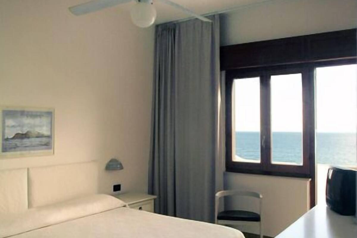 A hotel room with a bed, a desk, a chair, a ceiling fan, a window with a view of the sea, and a painting on the wall.