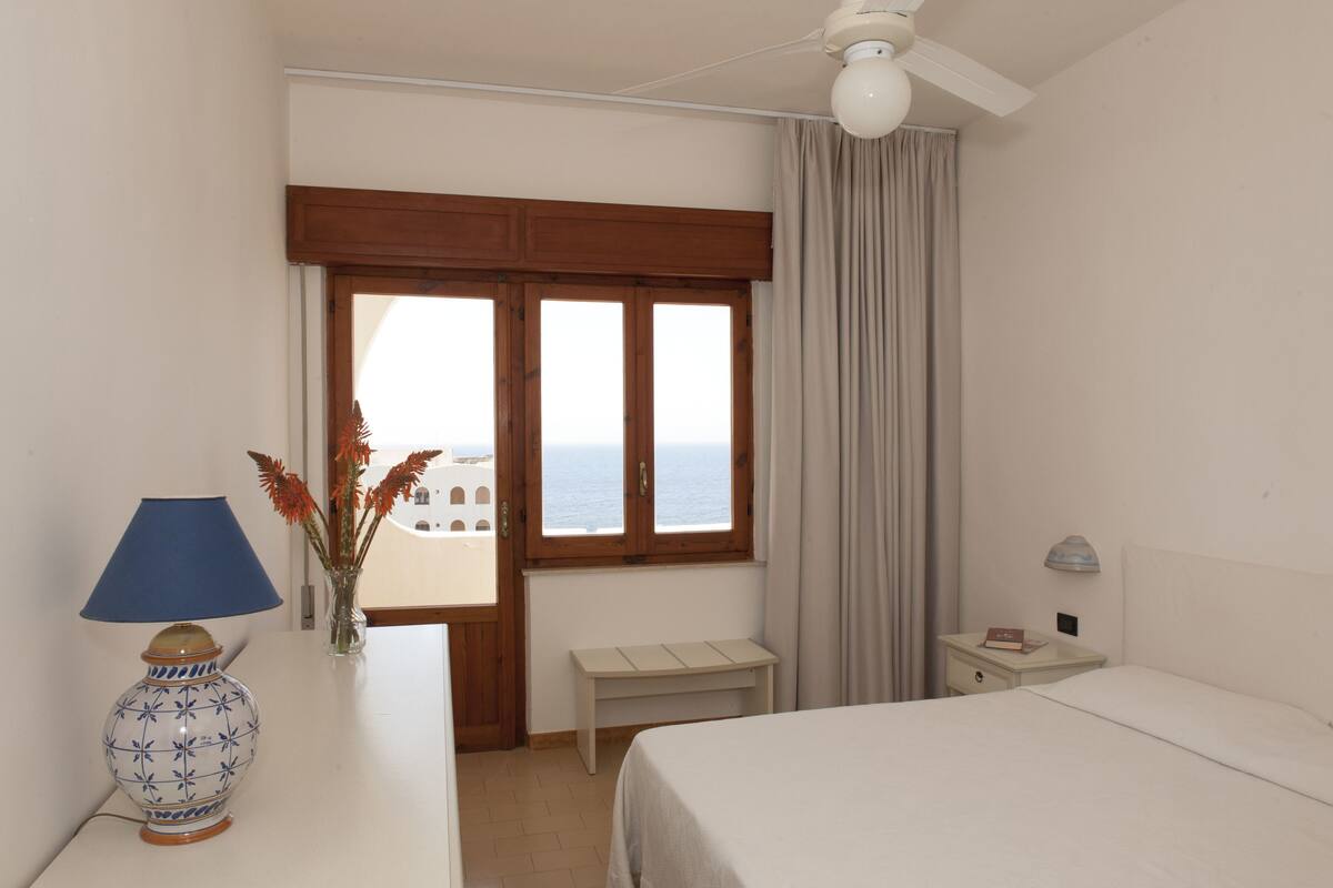A hotel room with a bed, a desk, a lamp, a vase with flowers, a window with curtains, and a ceiling fan.