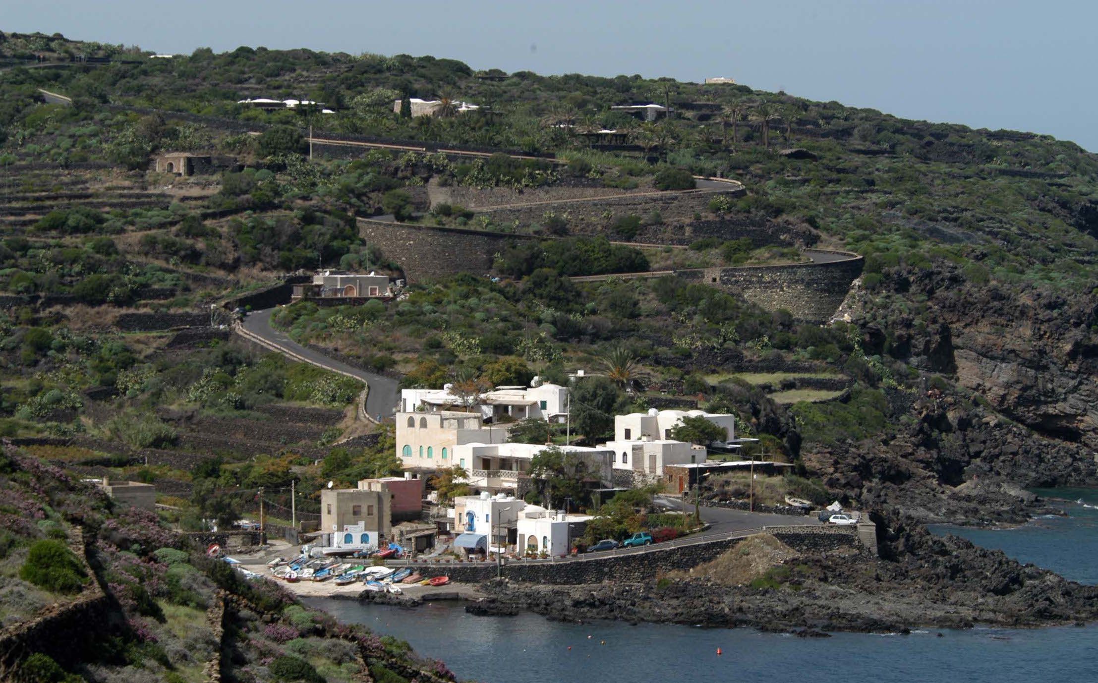 Uma vila costeira com casas em socalcos, uma estrada sinuosa e uma marina com barcos.
