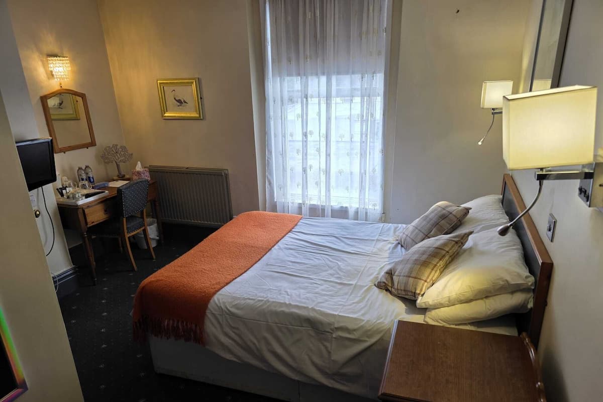 A hotel room with a bed, a desk, a chair, a radiator, a window with sheer curtains, and a wall-mounted lamp.