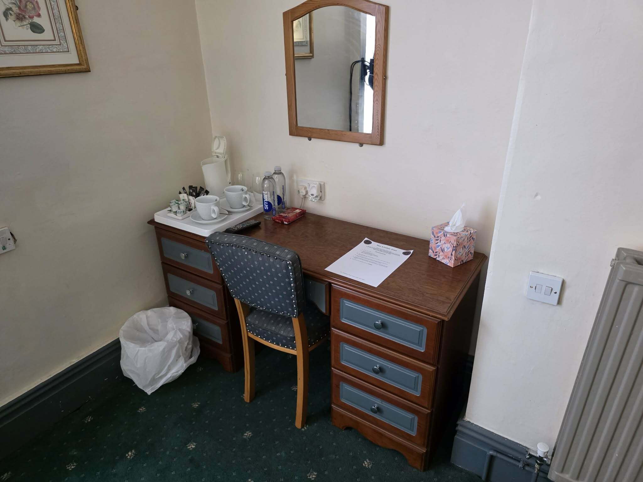 Une petite chambre d’hôtel avec un bureau en bois, une chaise, un miroir, un radiateur et une poubelle.