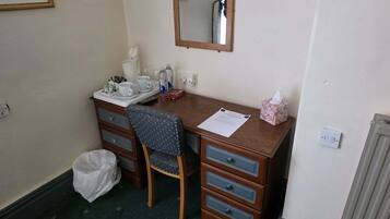 Une petite chambre d’hôtel avec un bureau en bois, une chaise, un miroir, un radiateur et une poubelle.