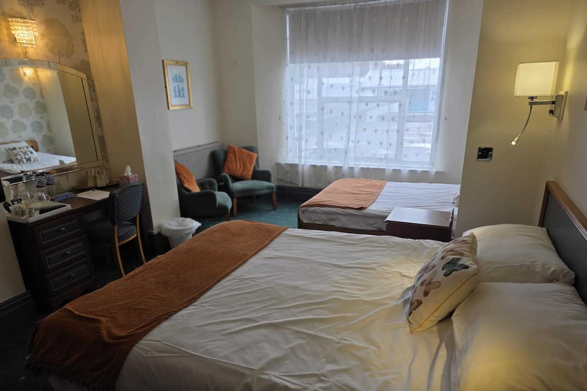 A hotel room with two beds, a desk with a mirror, a chair, a window with curtains, and a wall-mounted lamp.