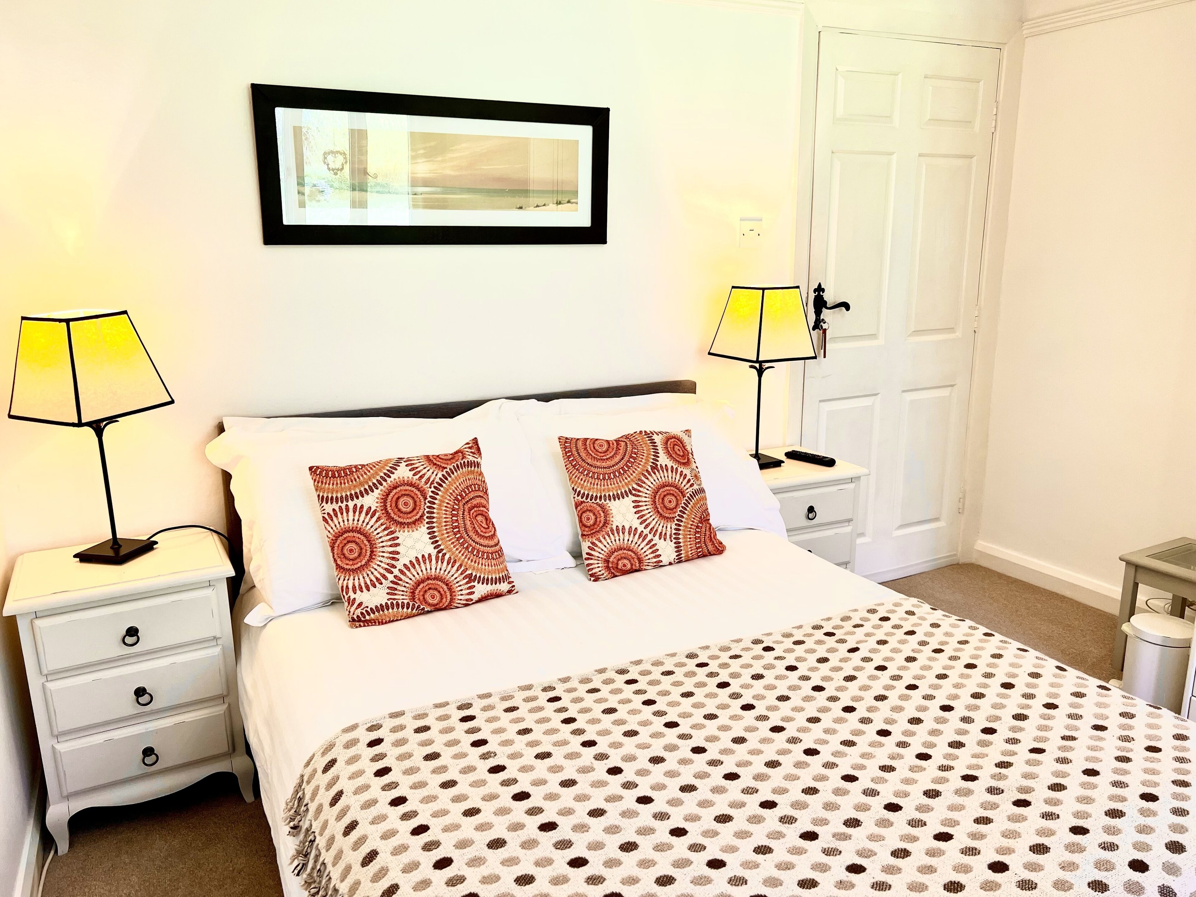 Une chambre bien rangée, avec un lit, deux tables de chevet, une lampe sur chacune, un tableau encadré au mur et une porte sur le côté droit.