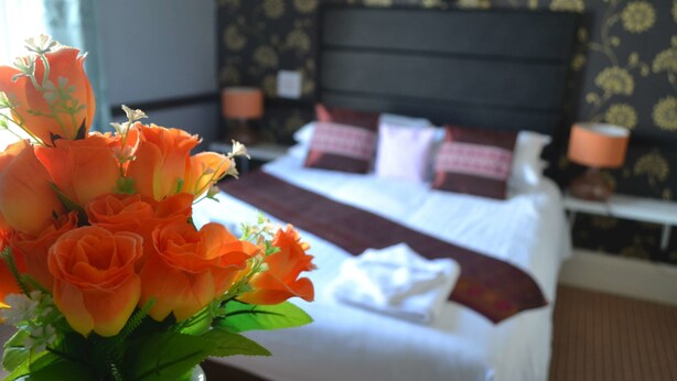 A hotel room with a neatly made bed, a vase of orange flowers, and a patterned wallpaper. - Holmlea Guest House (Blackpool)