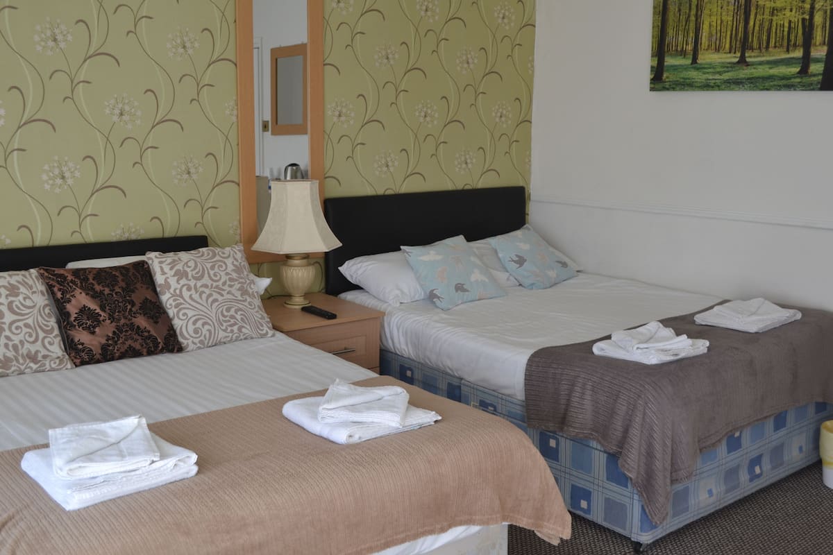 A hotel room with two beds, a nightstand, a lamp, and a painting of a forest on the wall.