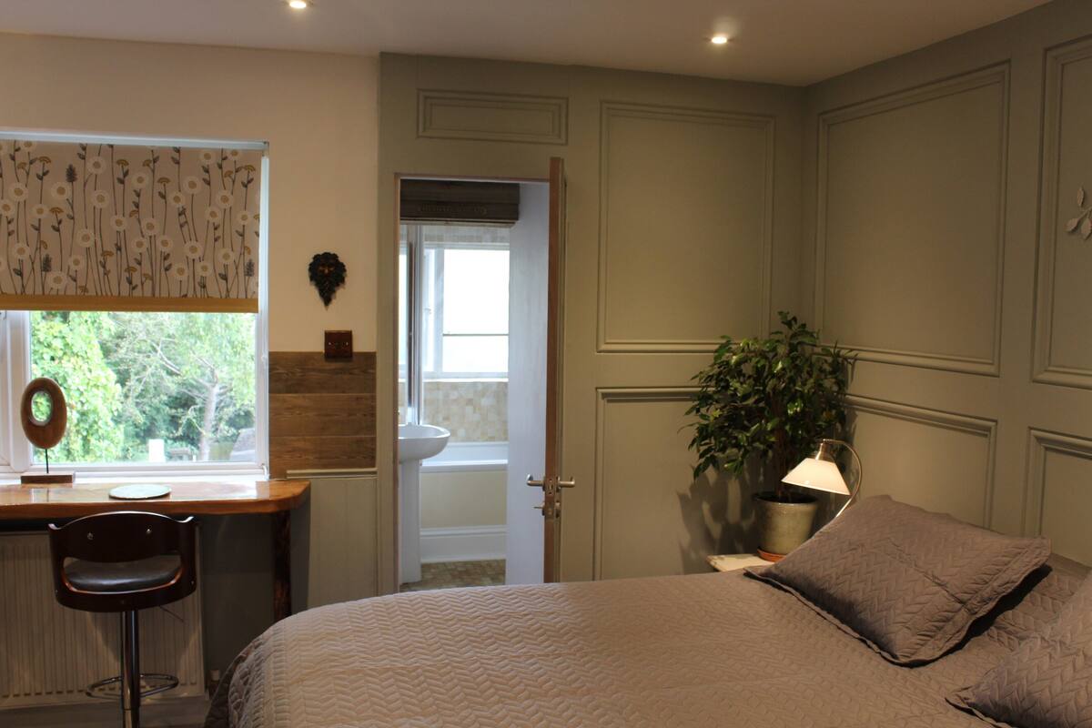 A bedroom with a bed, a desk, a chair, a potted plant, and a bathroom door.