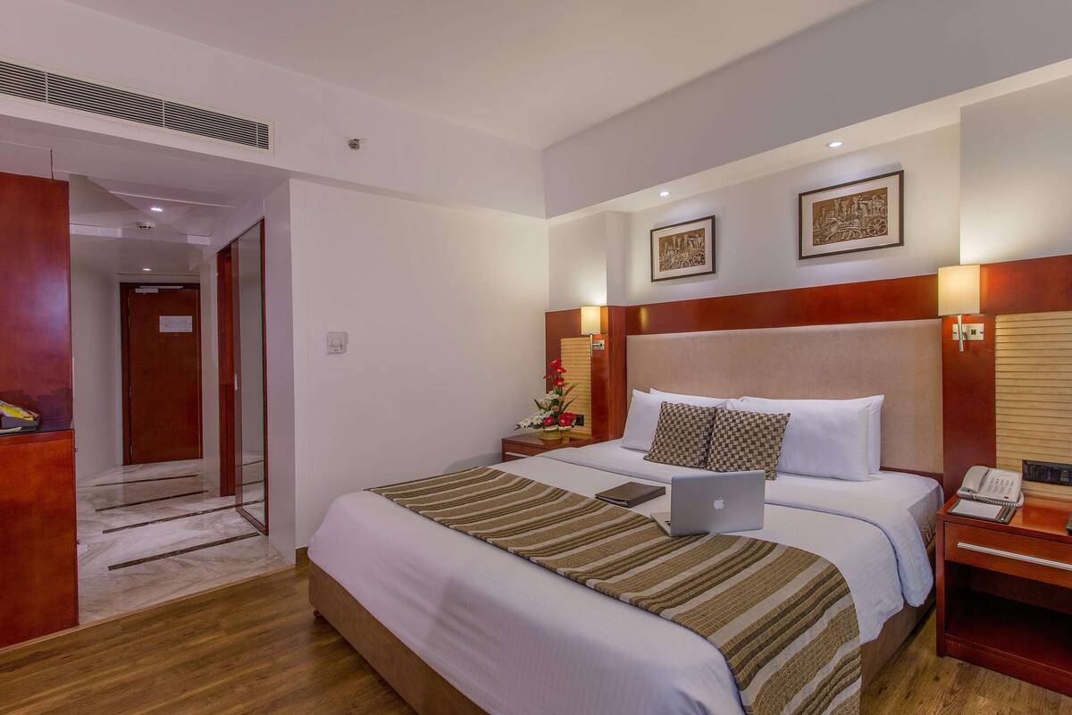 Une chambre d’hôtel avec un grand lit, un bureau avec un ordinateur portable, une table de chevet avec une lampe et deux tableaux encadrés au mur.