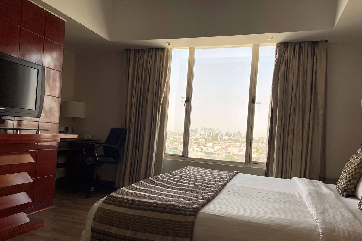 Une chambre d’hôtel dotée d’un grand lit, d’un téléviseur fixé au mur, d’un bureau et d’une chaise. La chambre offre une vue sur la ville grâce à de grandes fenêtres.