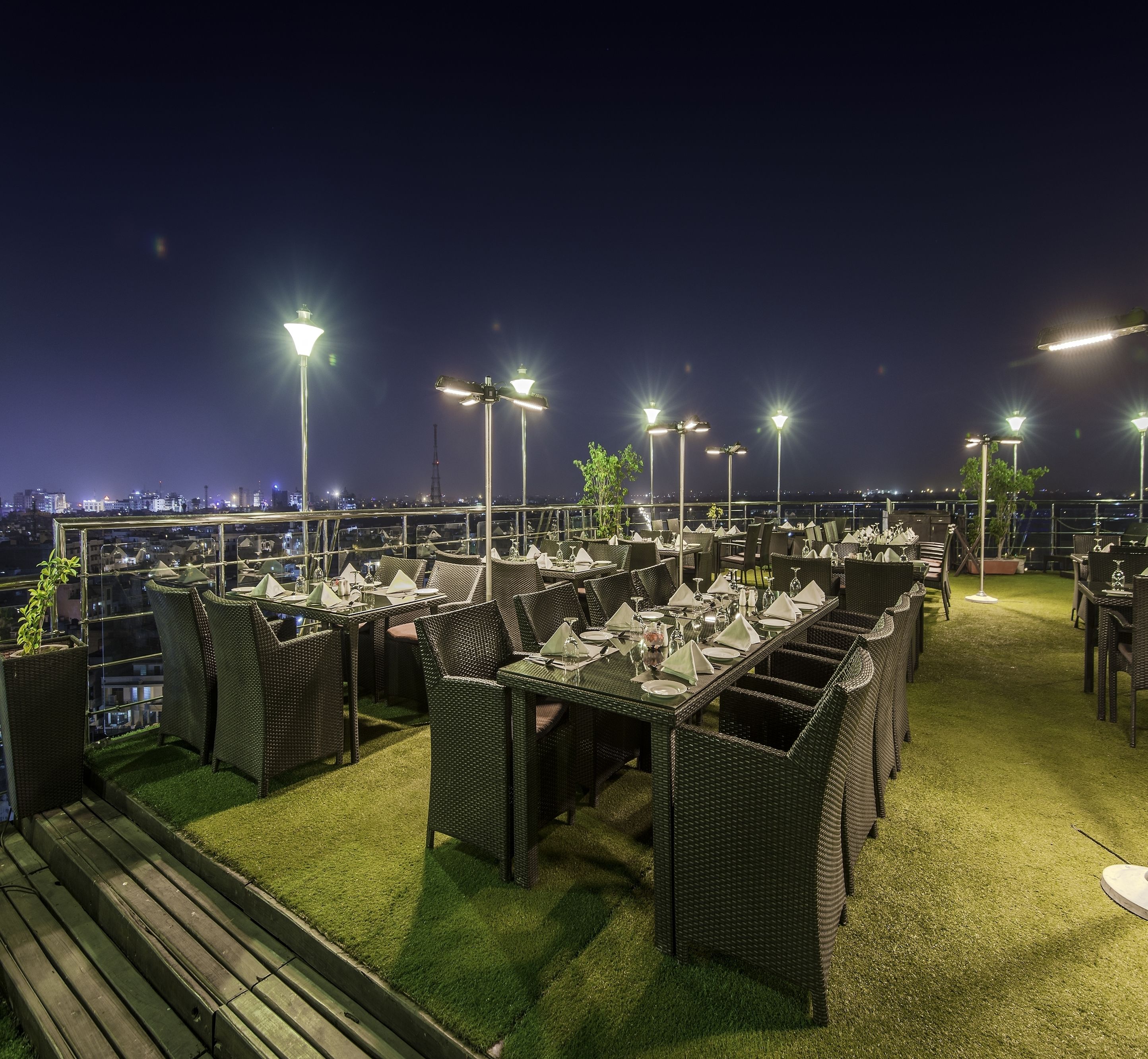 Un área de comedor al aire libre con varias mesas y sillas, rodeada de césped artificial e iluminada por farolas.