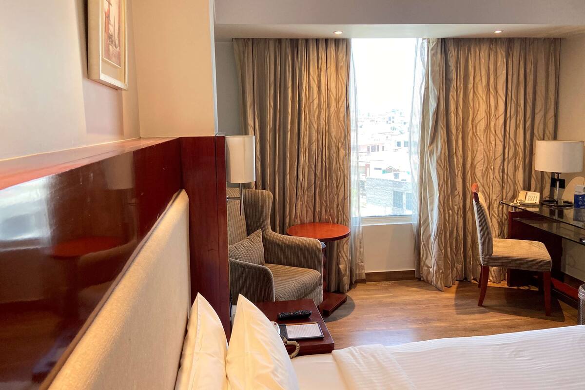 Une chambre d’hôtel avec un lit, un bureau doté d’une lampe, une chaise, une petite table et une vue sur la ville grâce à de grandes fenêtres équipées de rideaux.