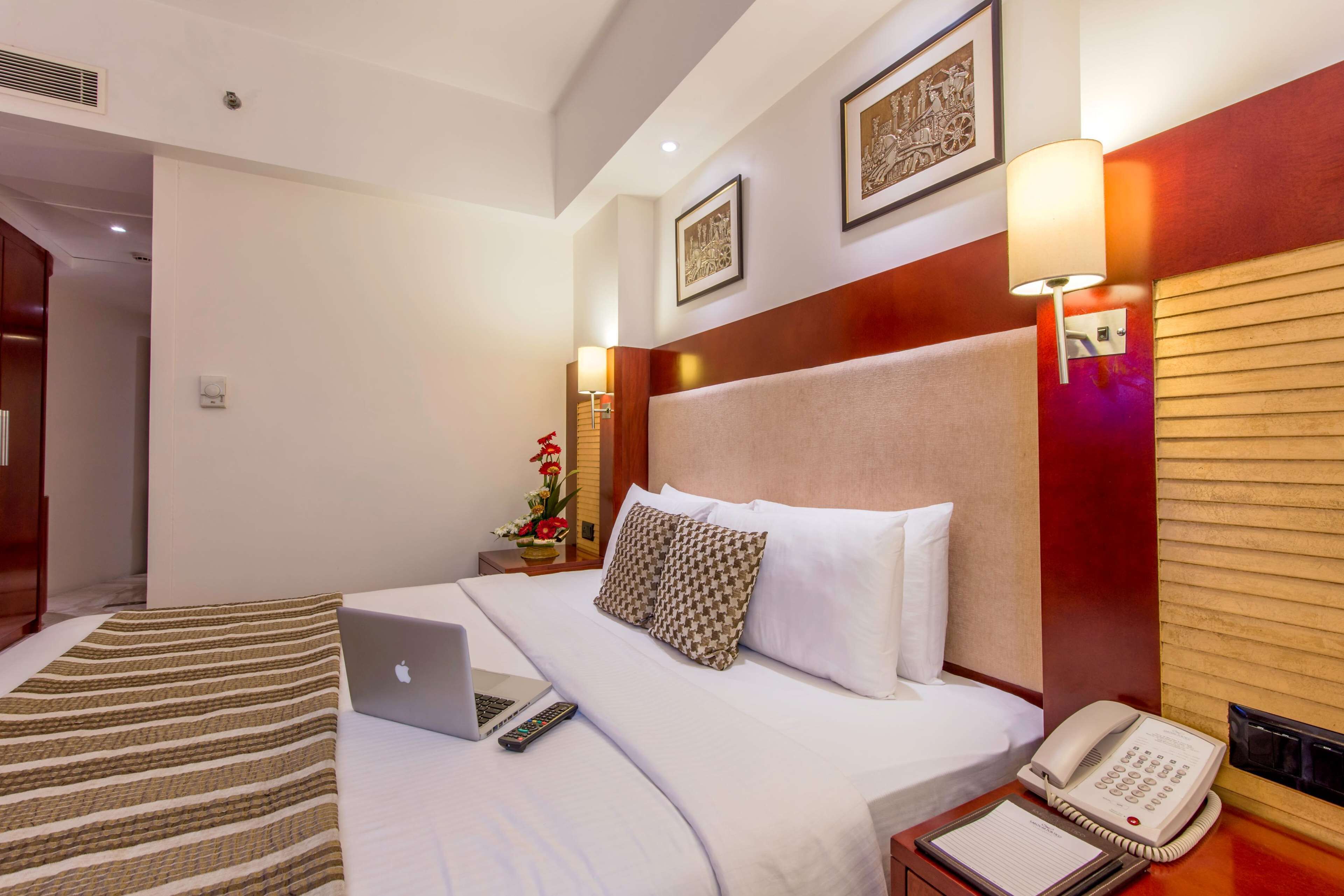 A hotel room with a bed, a desk with a laptop, a telephone, and a wall-mounted lamp.