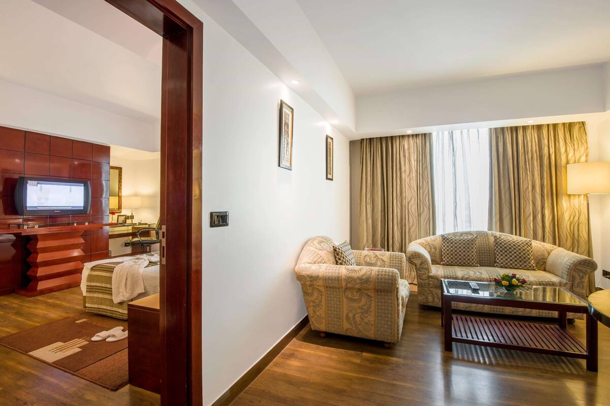 Une chambre d’hôtel dotée d’un espace salon comprenant un canapé, une table basse, un téléviseur fixé à un meuble en bois et une grande fenêtre avec des rideaux.