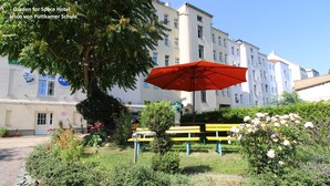 Um jardim com um guarda-sol vermelho, bancos e um edifício com um mural ao fundo.