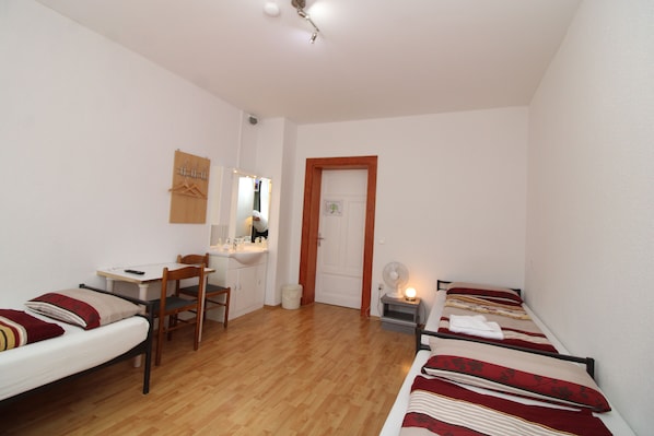 Una habitación pequeña y bien iluminada, con suelo de madera, una cama con ropa de cama roja y blanca, un escritorio con una silla, una mesita y un espejo.