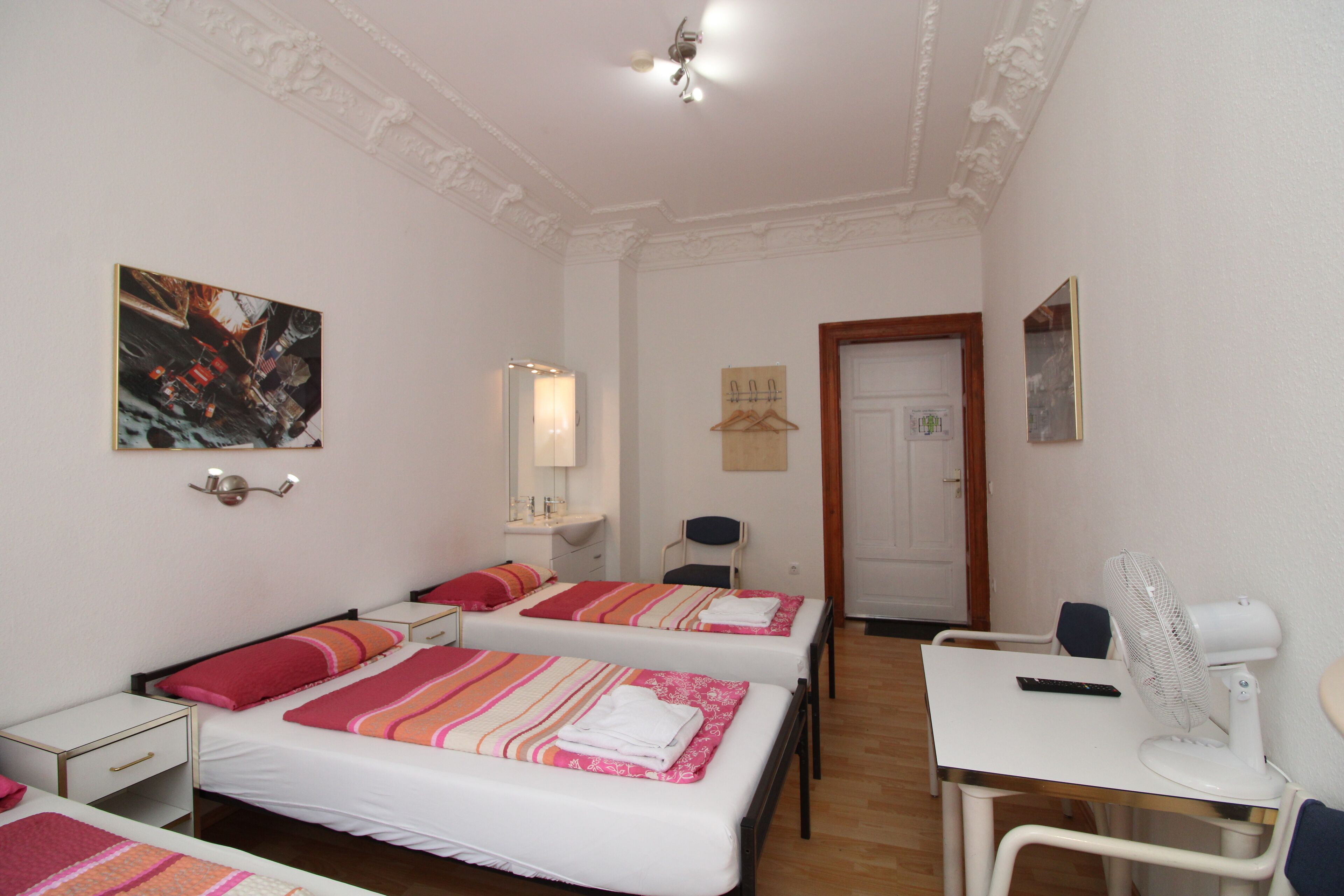 A hotel room with two beds, a desk, a chair, a fan, and a door.