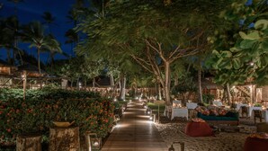 Un sentier en bois mène à une zone de plage aménagée avec des sièges et des tables, le tout entouré d’une végétation tropicale et de palmiers.