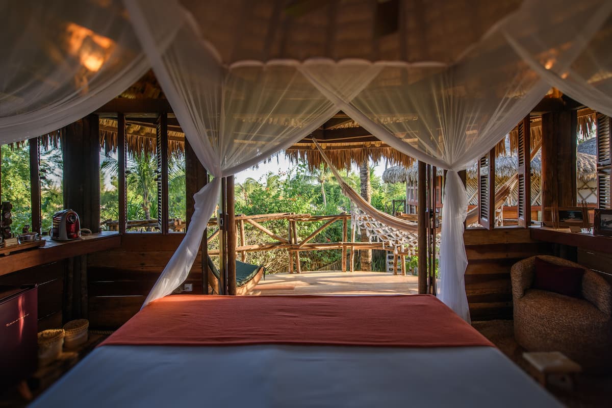 A room with a bed, a wooden desk, and a view of a wooden deck and palm trees.