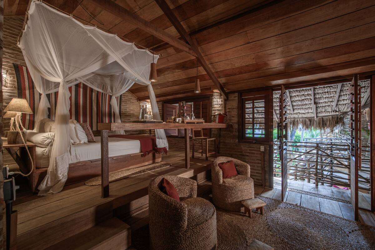 A rustic wooden room with a bed, a sitting area with woven chairs, a dining table, and a large window with a view of the outdoors.