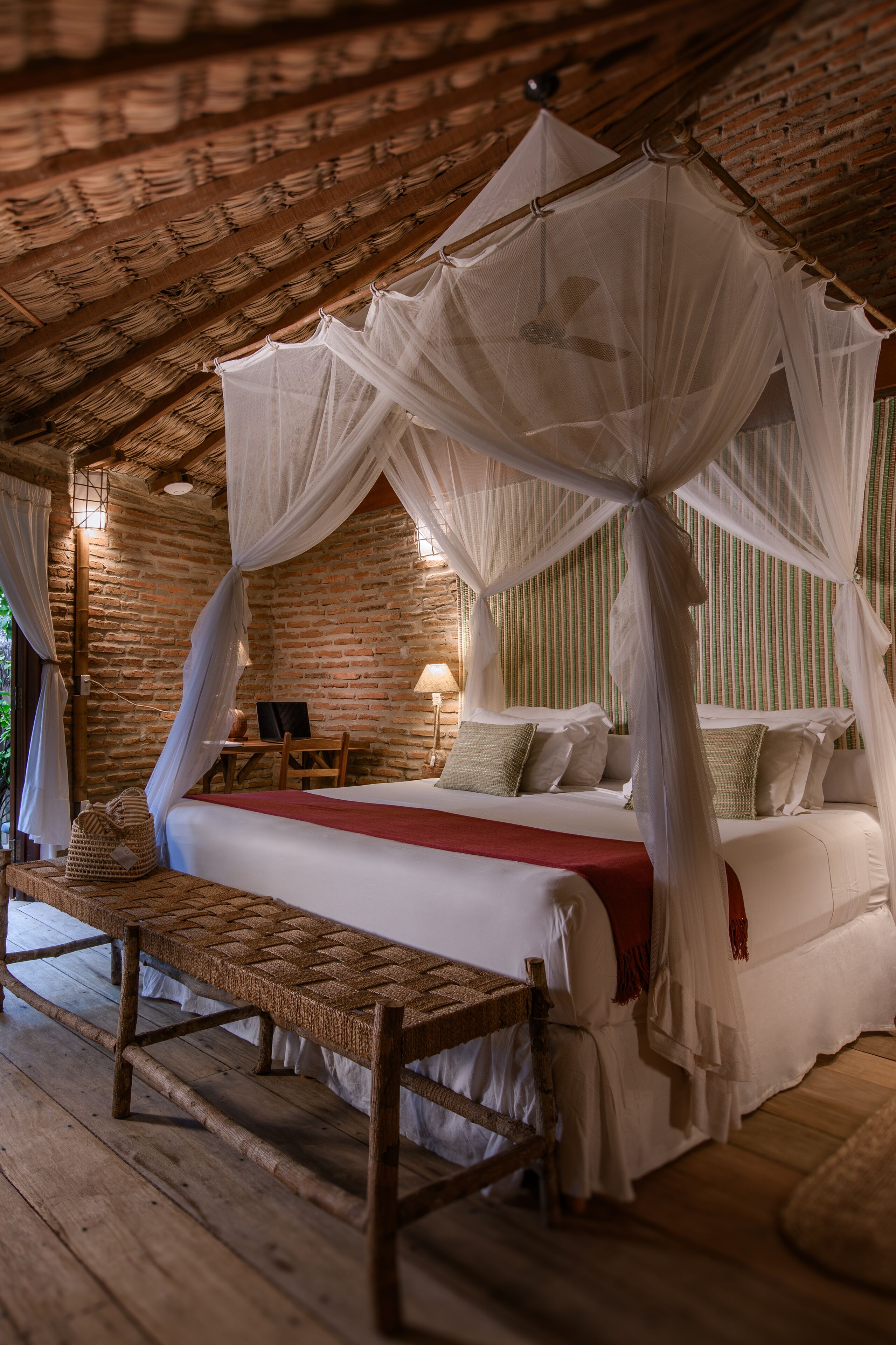 A rustic wooden room with a four-poster bed, a woven bench, and a brick wall.