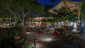 Un espace extérieur aménagé avec des tables et des chaises en bois, entouré d’une végétation luxuriante et bordé d’un bâtiment à toiture de chaume.