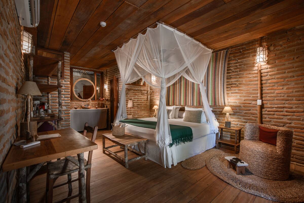A rustic room with a wooden ceiling, a four-poster bed with a canopy, a round mirror, a small table with a lamp, and a wicker chair.